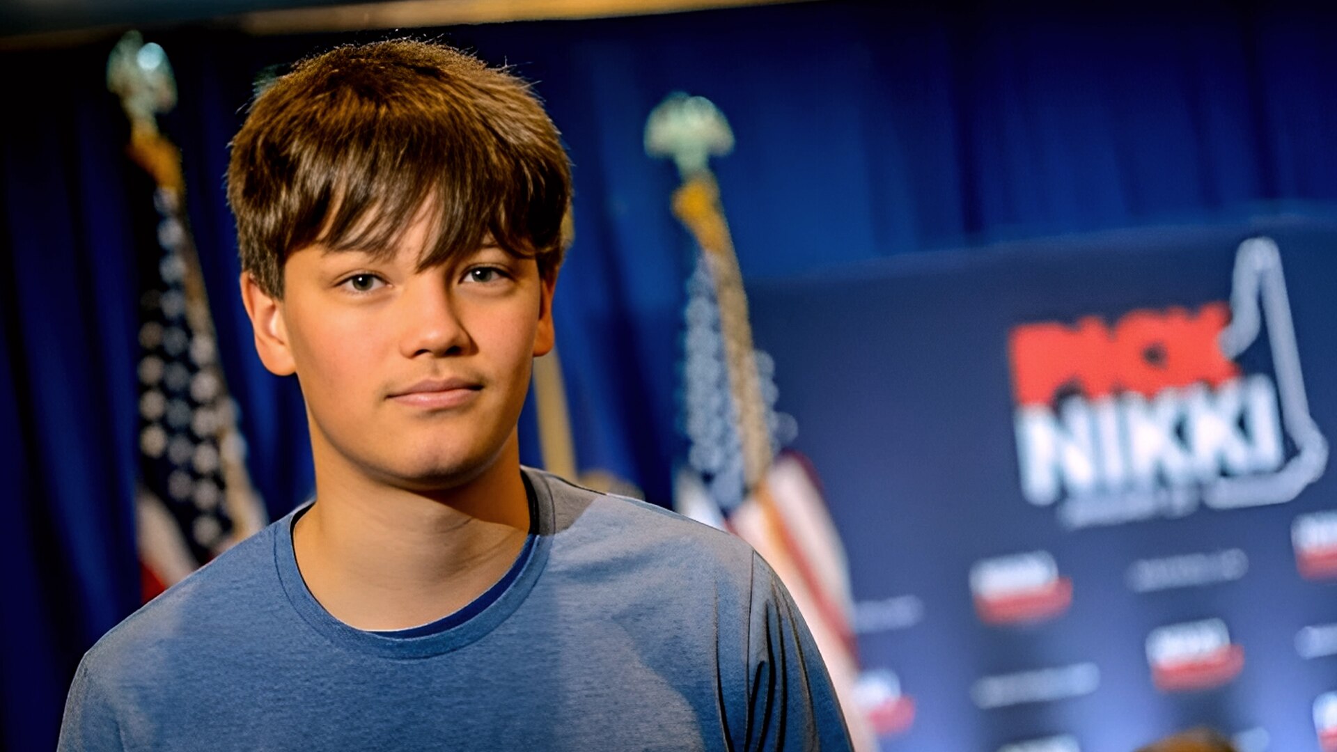 A young man in a blue sweater stands in front of blue stage, with US flags an a sign saying Pick Nikki. 