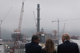 Three people stand overlooking a site with five cranes and structures being built.