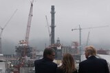 Three people stand overlooking a site with five cranes and structures being built.