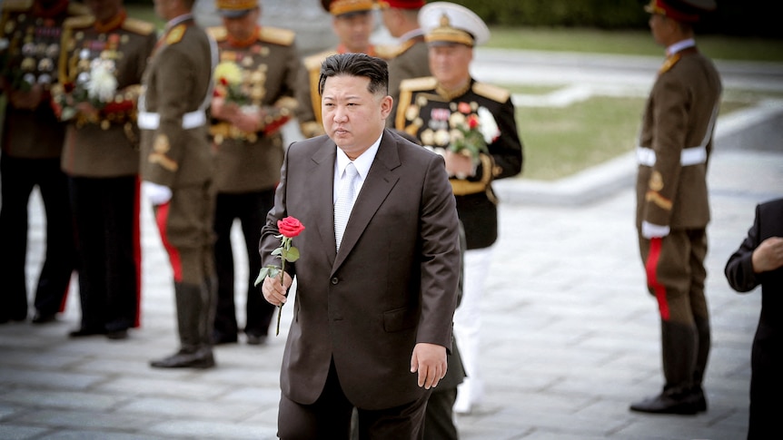 Kim Jong Un in a suit holding a red rose, with officers in dress uniforms behind him