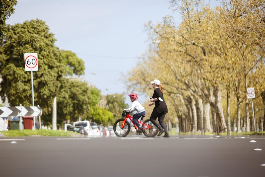 A woman running across a street next to a child on a bike.