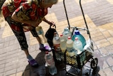An elderly woman leans over a cart filled with used plastic bottles containing water 