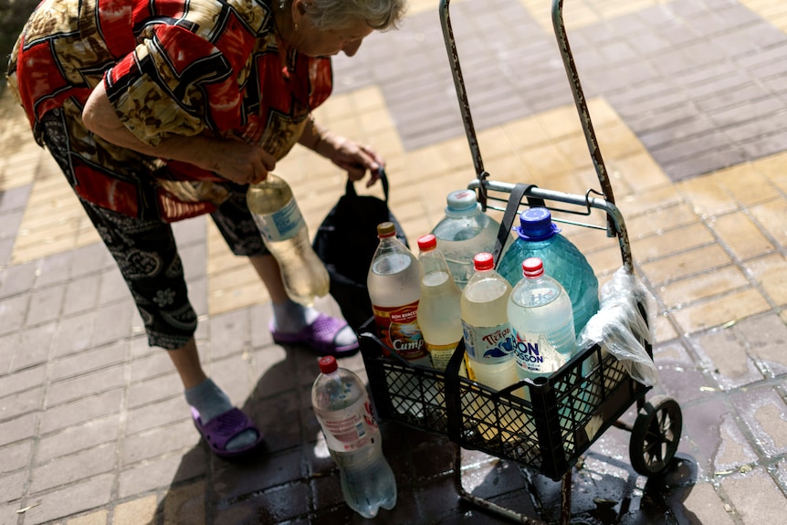 An elderly woman leans over a cart filled with used plastic bottles containing water 