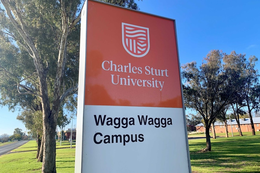 A red and white sign that has Charles Sturt University Wagga Wagga campus written on and is front of trees and a blue sky.