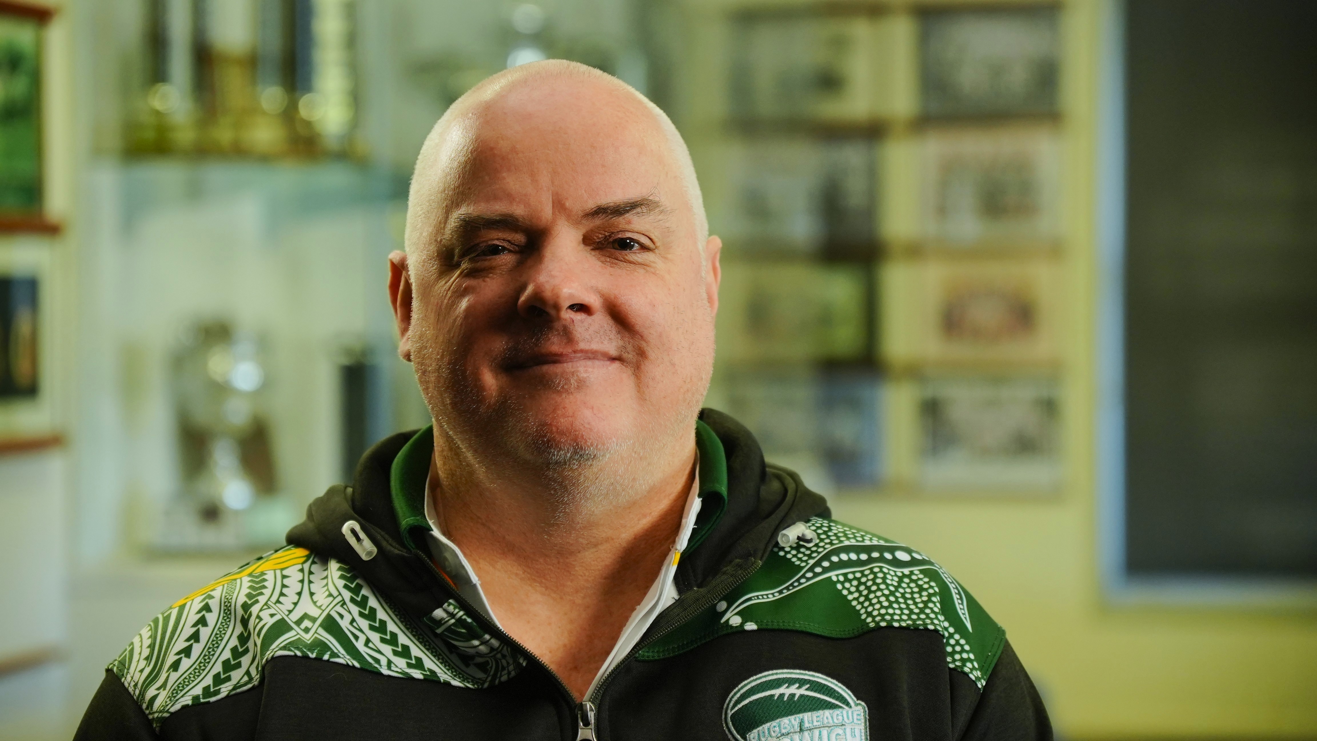 A bald man with white stubble poses for camera inside a room with cream coloured walls. There is a trophy cabinet behind him.