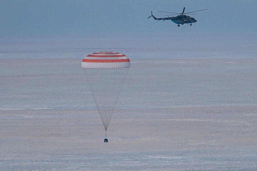 A capsule attached to a large parachute floats to the ground as a helicopter flies nearby.