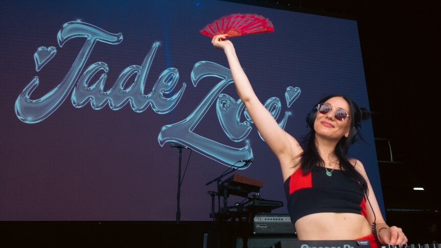 Jade Zoe DJing live in sunglasses and a black crop top, she's holding up a red fan and has her name logo behind her