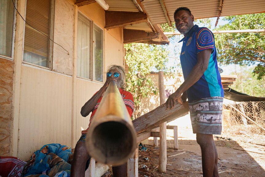 Djalu Gurruwiwi and his son Larry Gurruwiwi standing outside their home in Birritjimi holding didgeridoos.