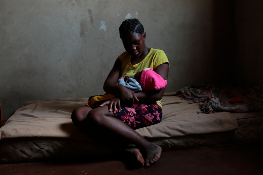 A young girl sits on the edge of a mattress on the ground holding a three-month-old child.