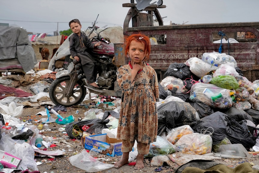 一个橘色头发的女孩赤着脚站在垃圾堆中。 她身后是一个男孩靠在摩托车上。 