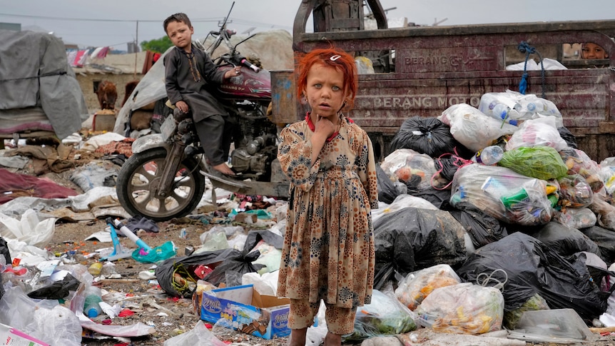 A girl with orange hair stands among rubbish with bare feet. A boys is behind her, leaning against a motorbike. 