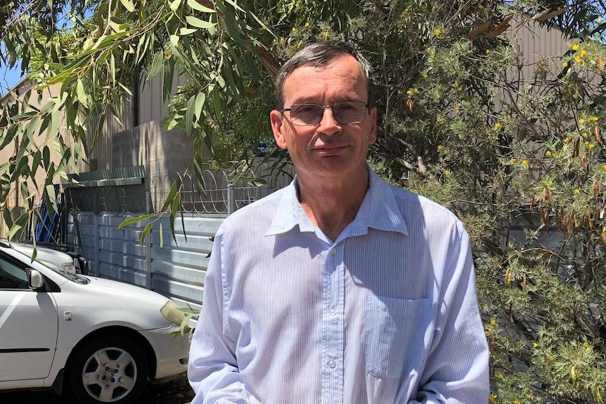 A man with grey hair and glasses stands in a carpark with trees in the background.