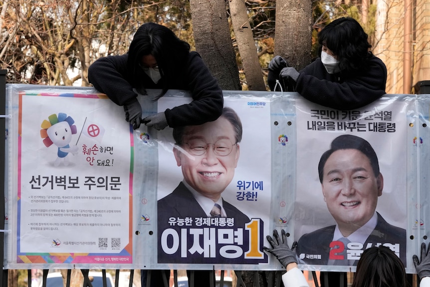 Masked people post election campaign posters featuring smiling middle-aged East Asian men