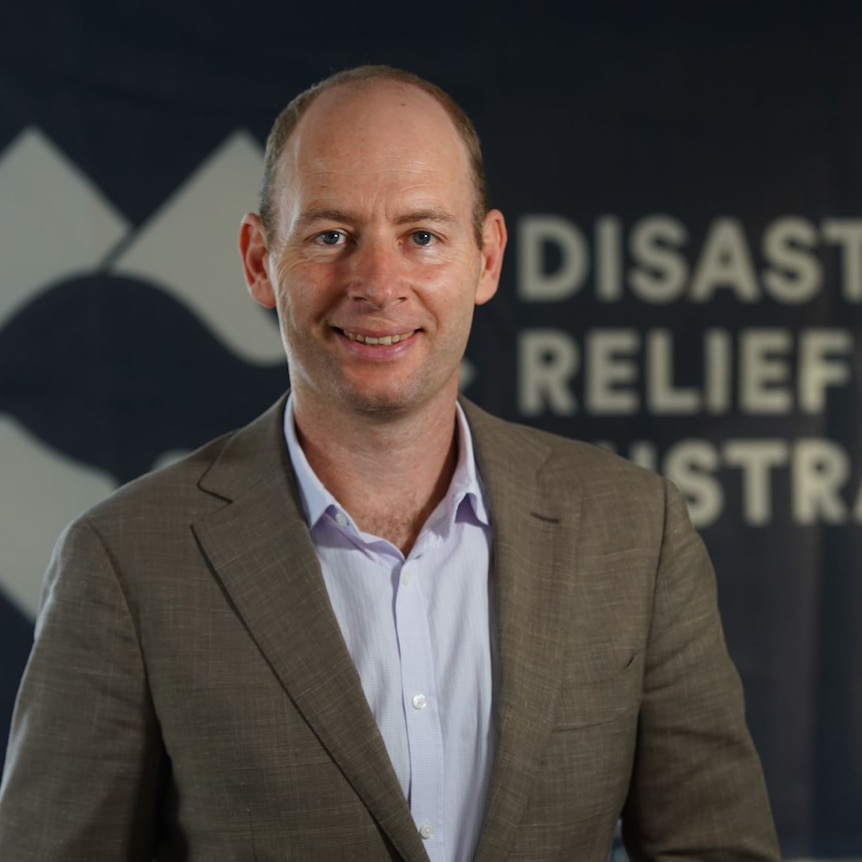 A man in suit with short cropped hair smiles at camera, standing in front of Disaster Relief banner.