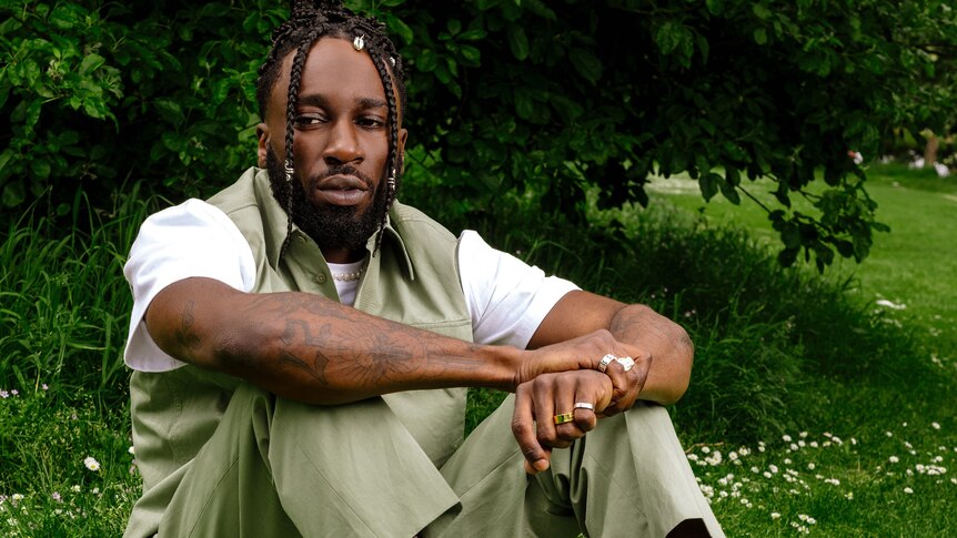 Kojey Radical sits outside on the grass wearing beige waistcoat, pants and white shirt