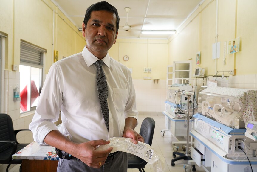 A man wearing a white shirt and tie stands in a hospital room holding a paper.