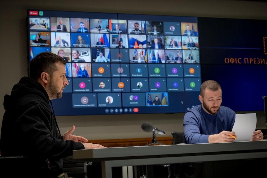 Two men sitting in front of a screen with an online meeting occuring
