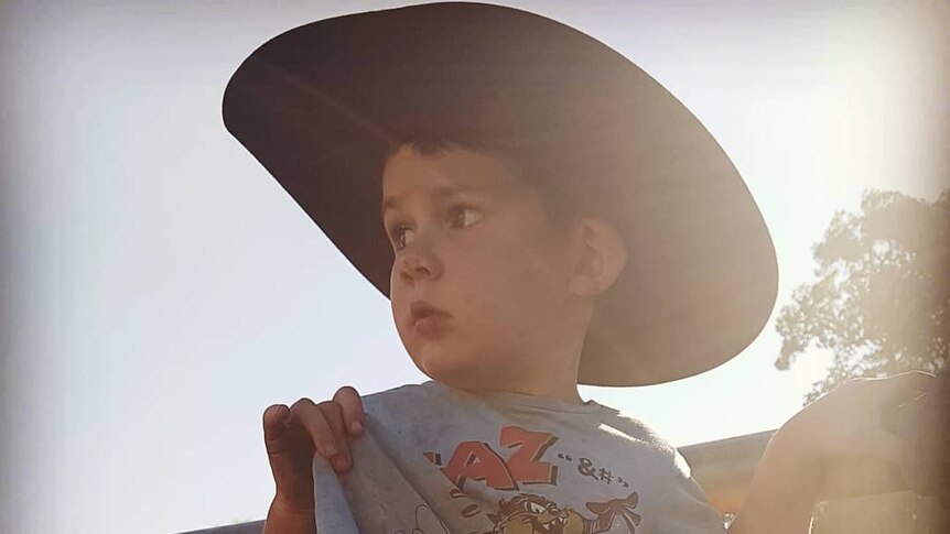 A young boy wearing a big hat stands beside cattle yards.