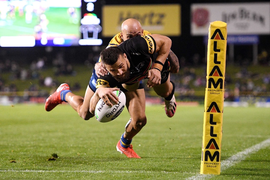 A Penrith NRL player dives in the air holding the ball with his right hand as he attempts to score a try while being tackled.