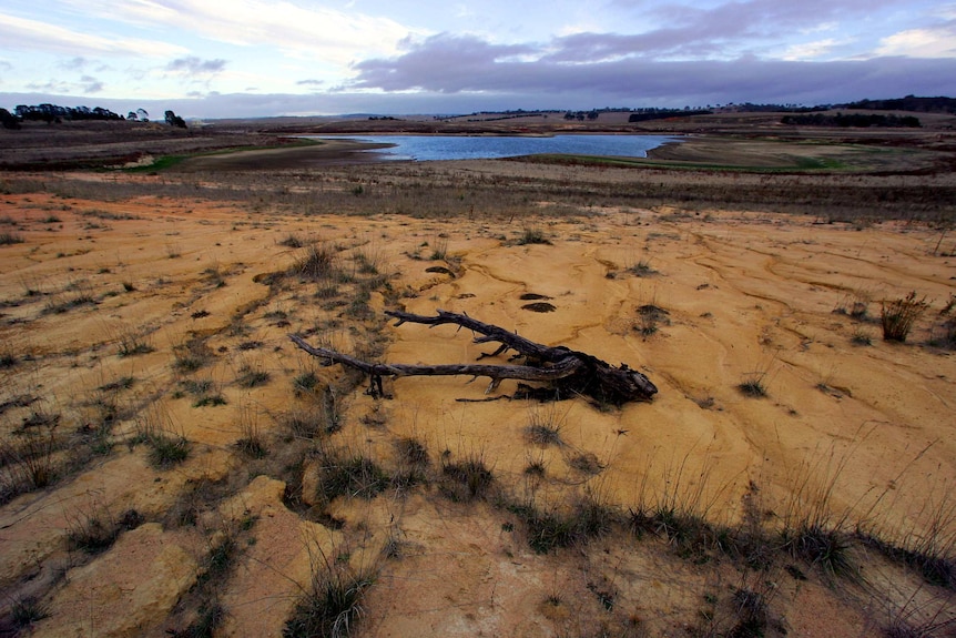 Sandy ground with patches of dry grass lead up to a large dam. The sky is cloudy