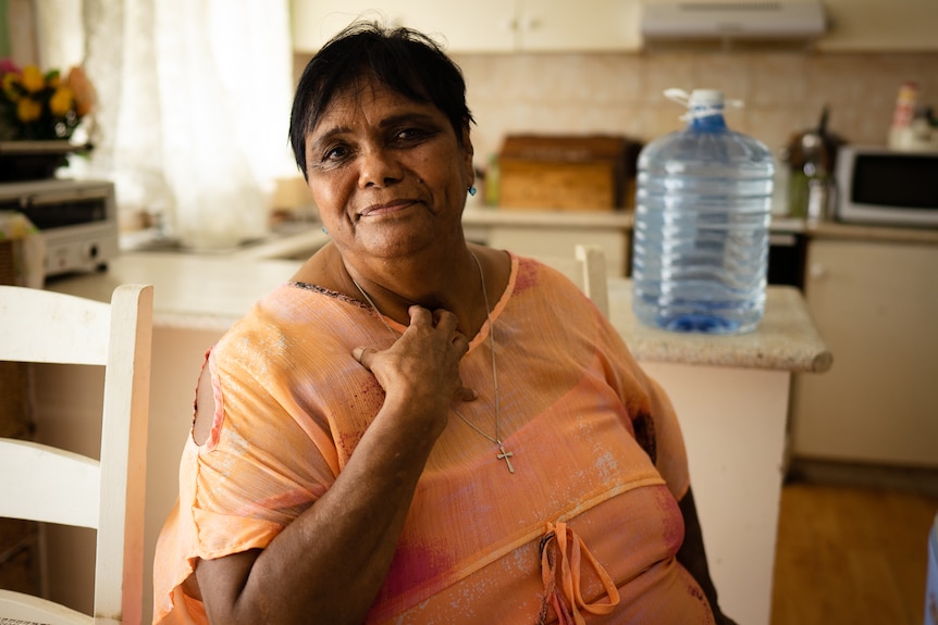 Indigenous woman wearing an orange top with her hand to her chest. A large bottle of water is behind her.