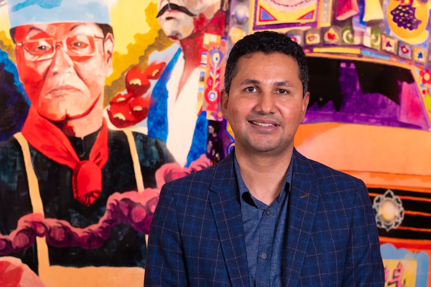 A man stands in front of a mural made up of Pakistani cultural references.