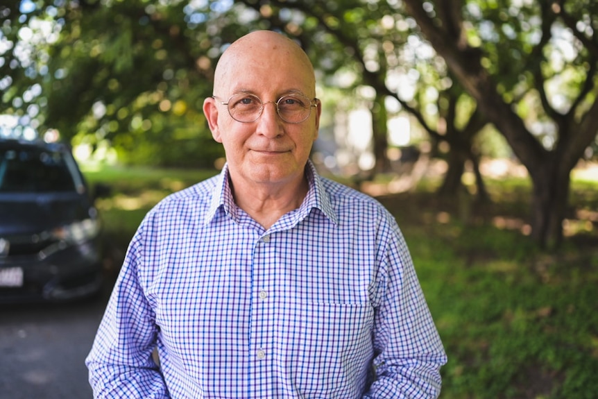 A bald, bespectacled man in a button-up shirt standing in front of some trees.