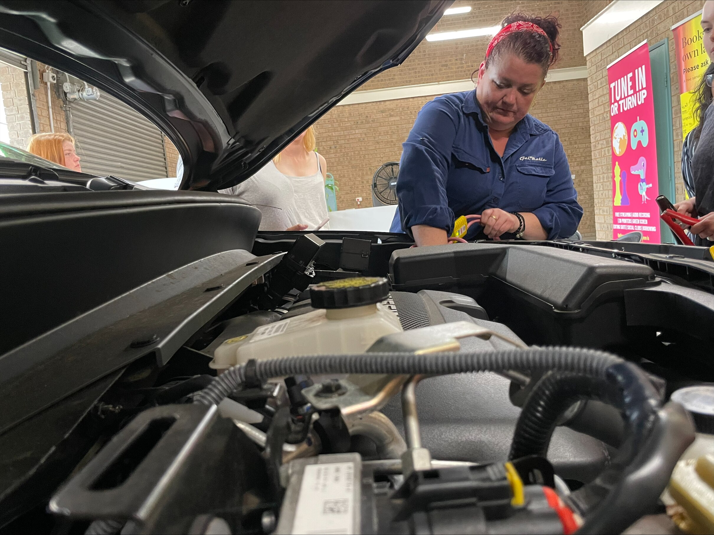 A woman in a blue denim shirt with her hand in a car engine.