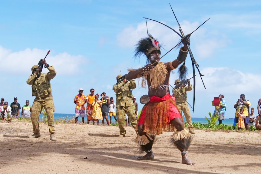 Torres Strait Islander dancer, dances out the front with bow & arrow,. In background dancers in ADF uniform dance with guns.