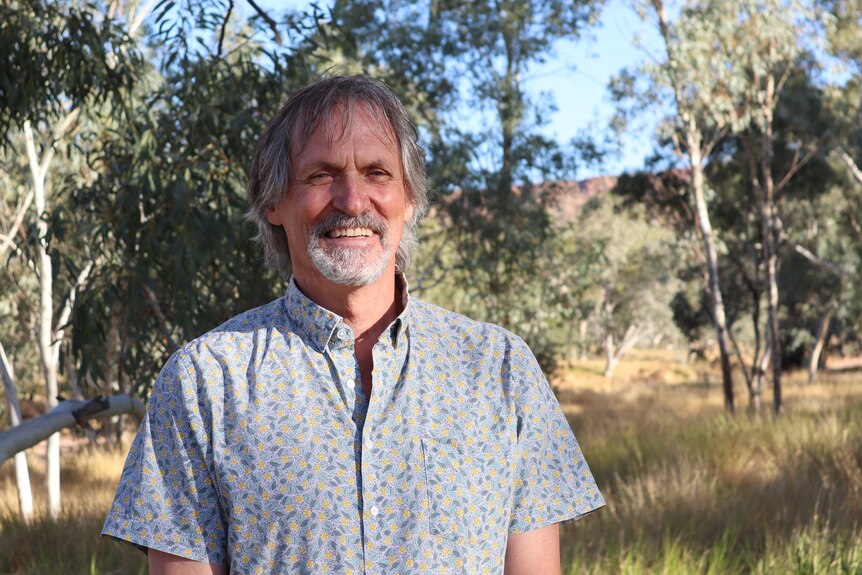 A man with a patterned shirt smiles as he stands with bushland in the background.