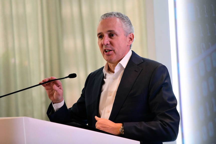 Telstra chief executive Andy Penn wears a black suit and speaks at a lectern.