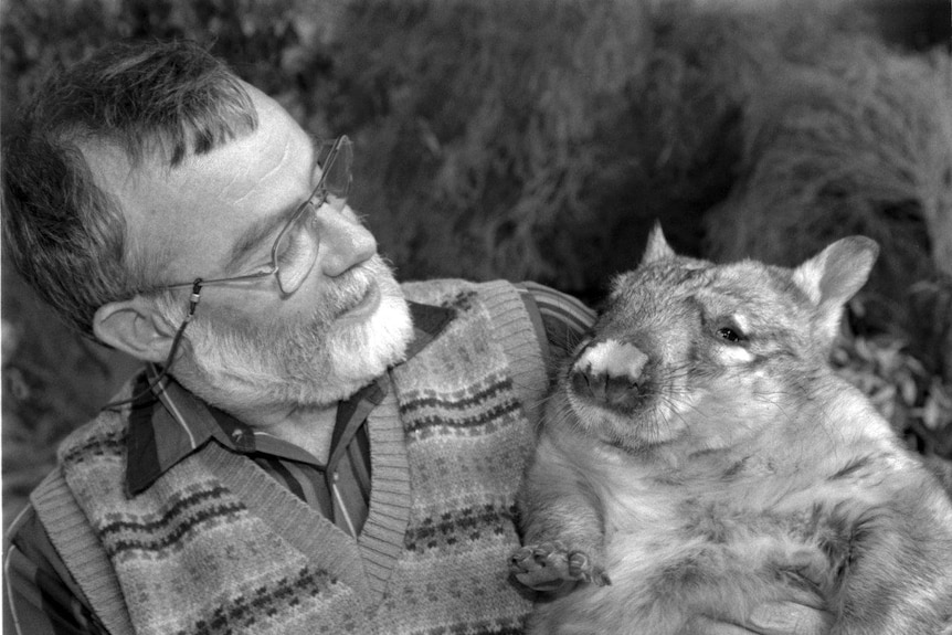 An older man with beard and glasses and a wearing a vest sits with a wombat in his left arm like they're buddies