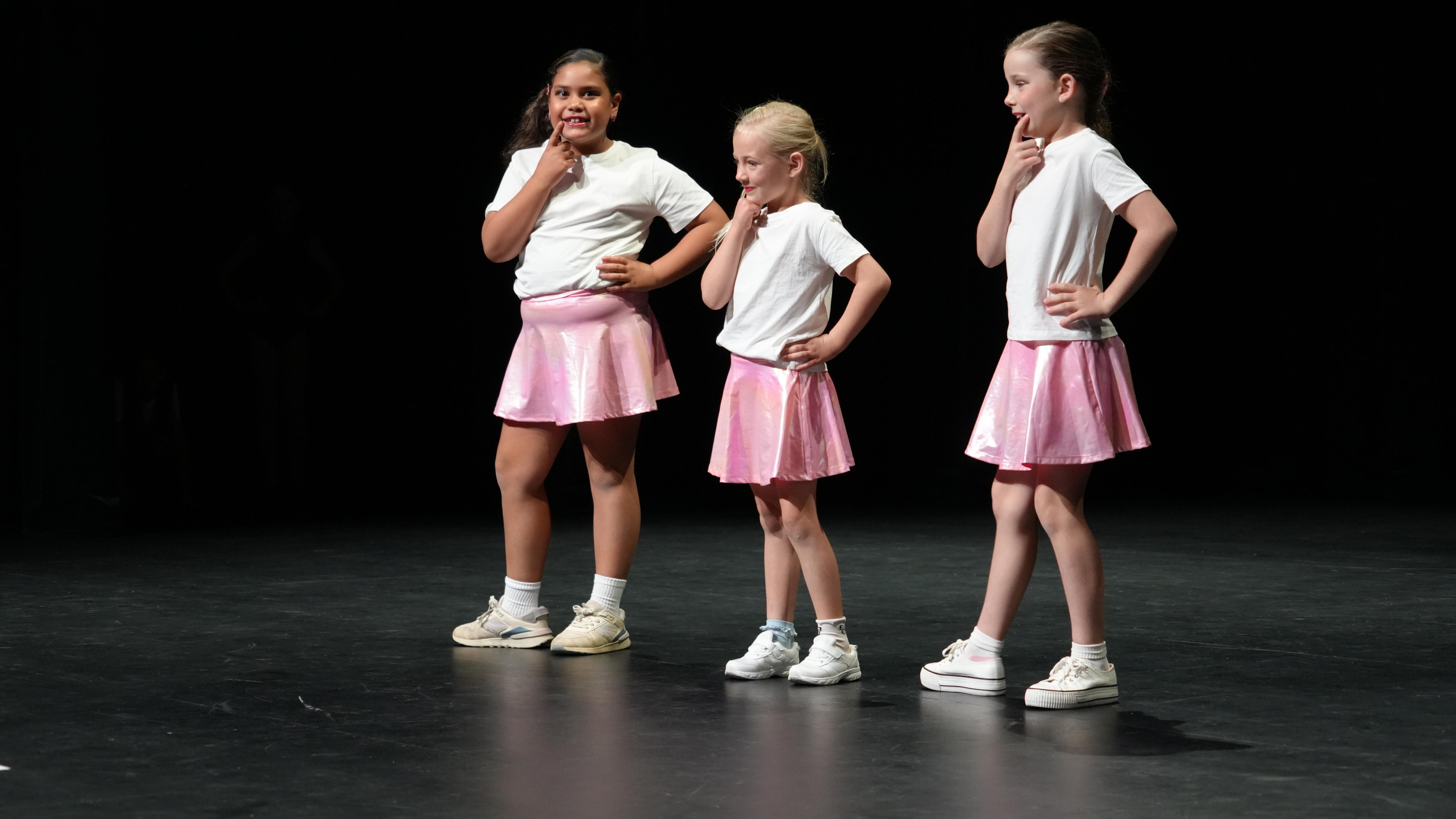 Tres chicas con camisas blancas y faldas de color rosa brillante posan con las manos en la barbilla en un escenario negro.