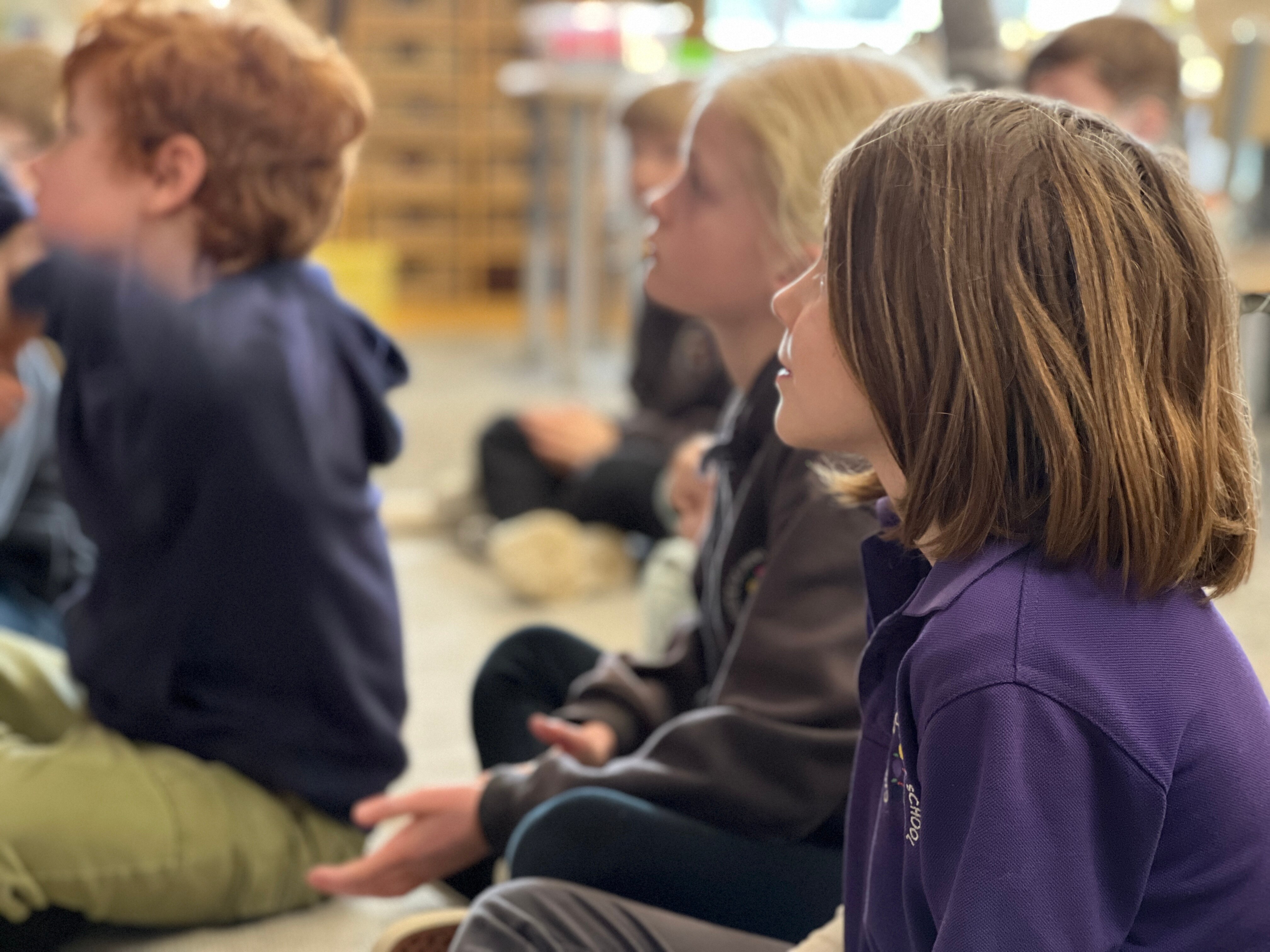 A girl with medium-length hair sitting on the floor with other children in the background wearing school uniform