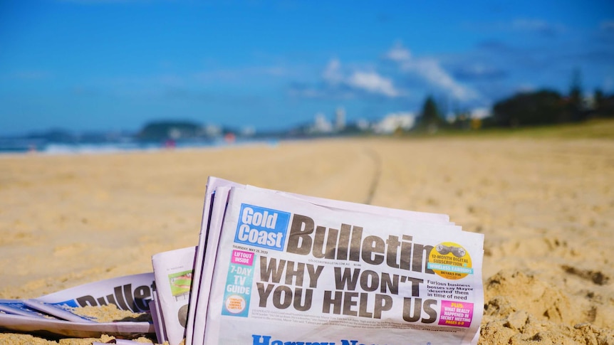 A newspaper – the Gold Coast Bulletin – sits in the golden sand of a sun-drenched beach.