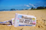 A newspaper – the Gold Coast Bulletin – sits in the golden sand of a sun-drenched beach.