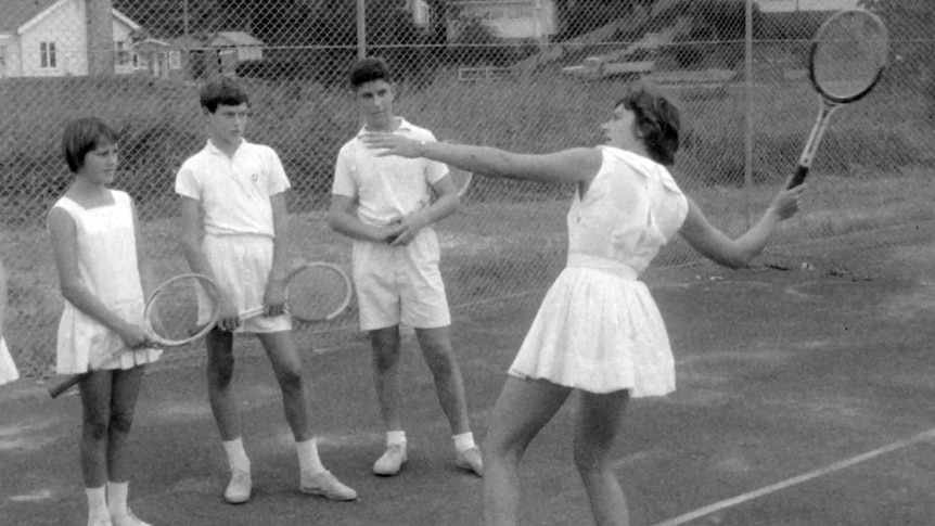Old photo of women demonstrating tennis shot in front of line of teenagers who watch