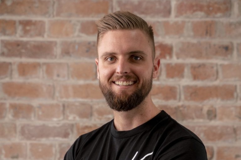 A young man with short hair and a beard stands in front of a brick wall and smiles.