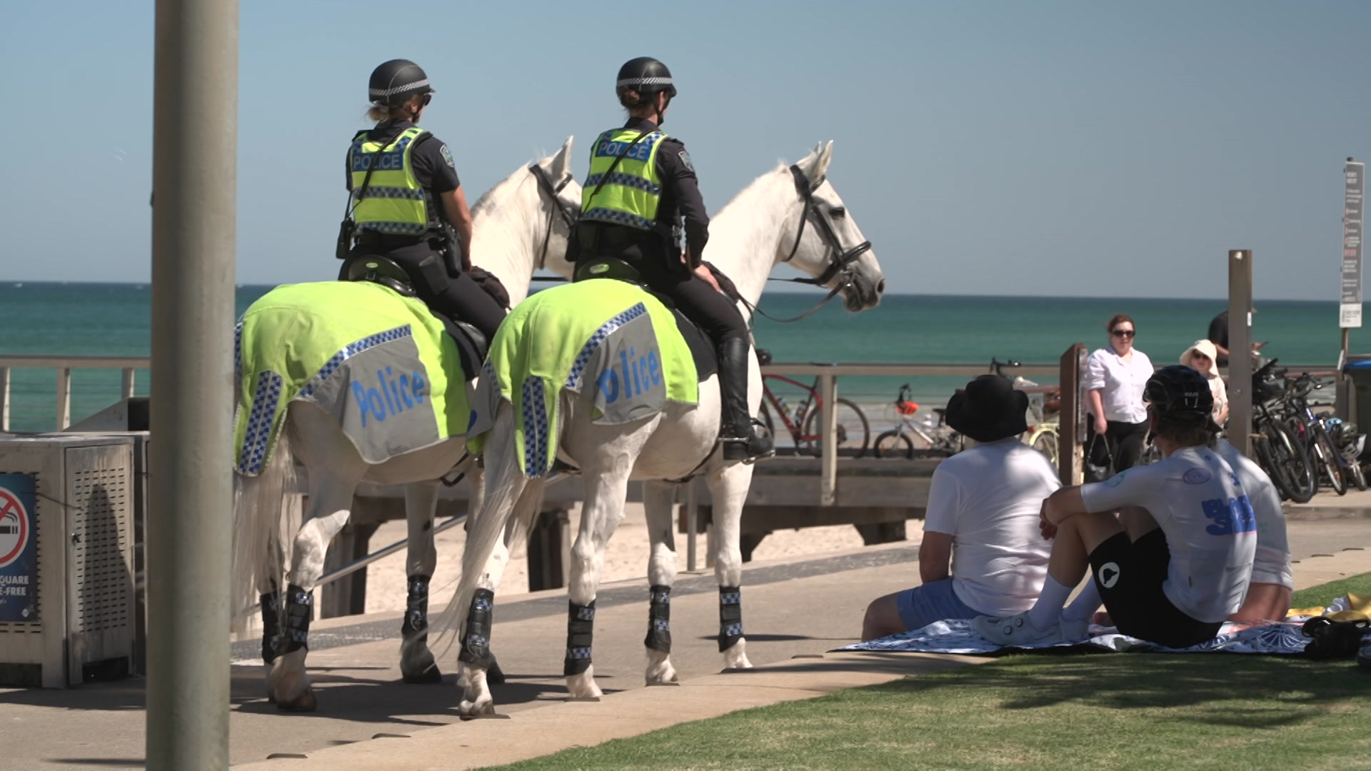 Dois policiais montando cavalos brancos na praia com outras pessoas ao redor