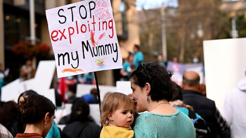 A woman in a green jumper holds a baby in a yellow jumper while holding a protest sign.