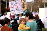 A woman in a green jumper holds a baby in a yellow jumper while holding a protest sign.