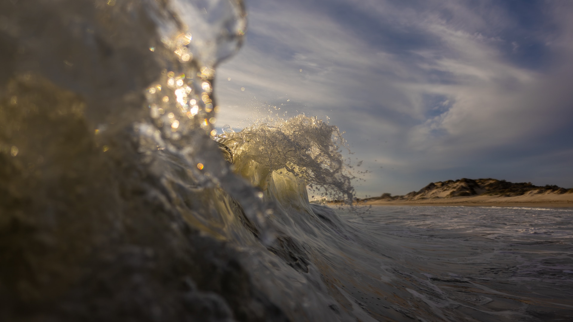 A in the water photo of a wave crashing near shore.  There are clouds in the sky and it's late afternoon golden hour.