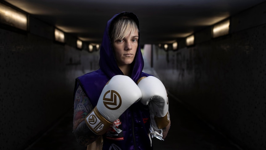 A young tattooed woman with boxing gloves on stares at the camera.