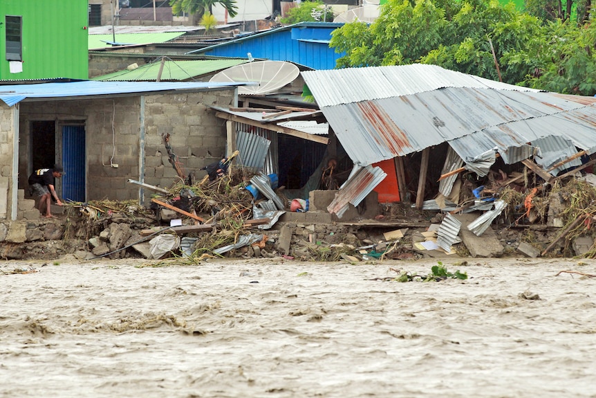 A man inspects the wreckage of buildings damaged by a flood, with surging brown floodwaters only metres from the structures.