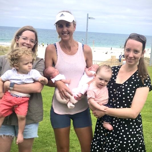 Three ladies standing infront of beach, holding their babies in their arms.