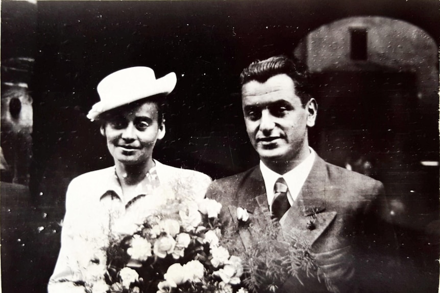 An old photo of a man and woman on their wedding day. Woman is wearing a white hat and holding a bunch of white roses.
