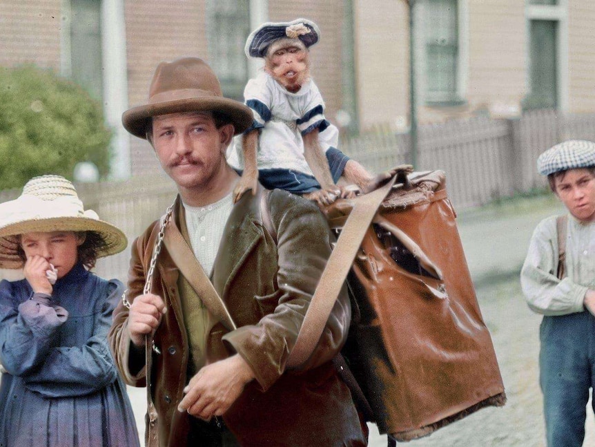 A man surrounded by children has a dressed monkey on his large carry bag.