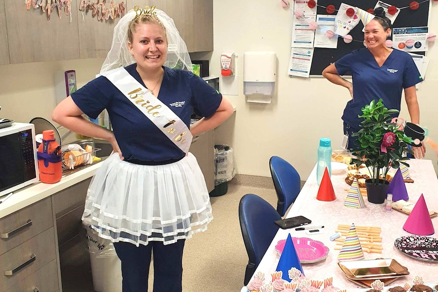 A woman stands next to a table of food wearing a tutu, sash and veil over her nurse's uniform.