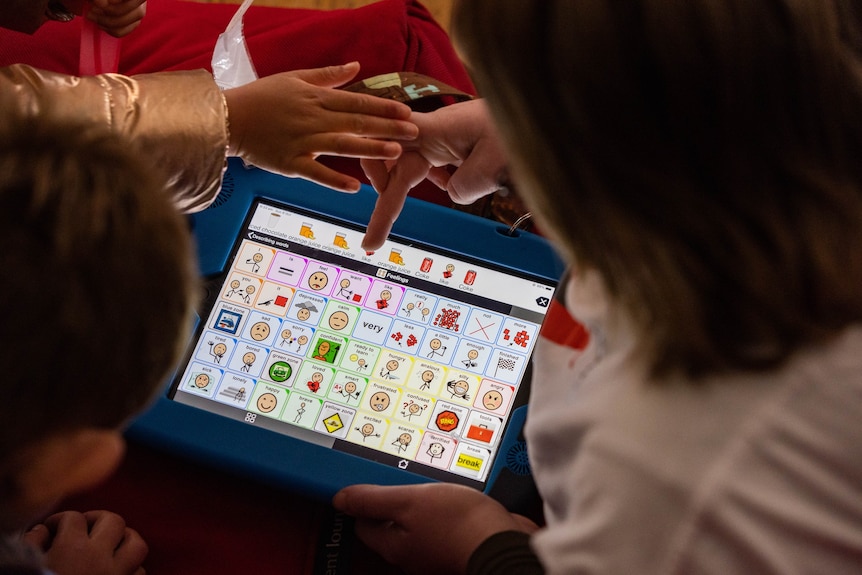 Looking over the shoulders of two children pointing to a laptop-sized device displaying a grid of images
