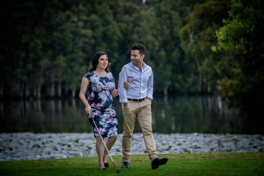 Nas and Tom walking together across a lawn in front of a lake surrounded by trees, Tom is looking at Nas while she speaks.